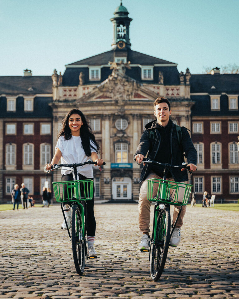 Young woman and a young man smiling and riding green bicycles on the campus